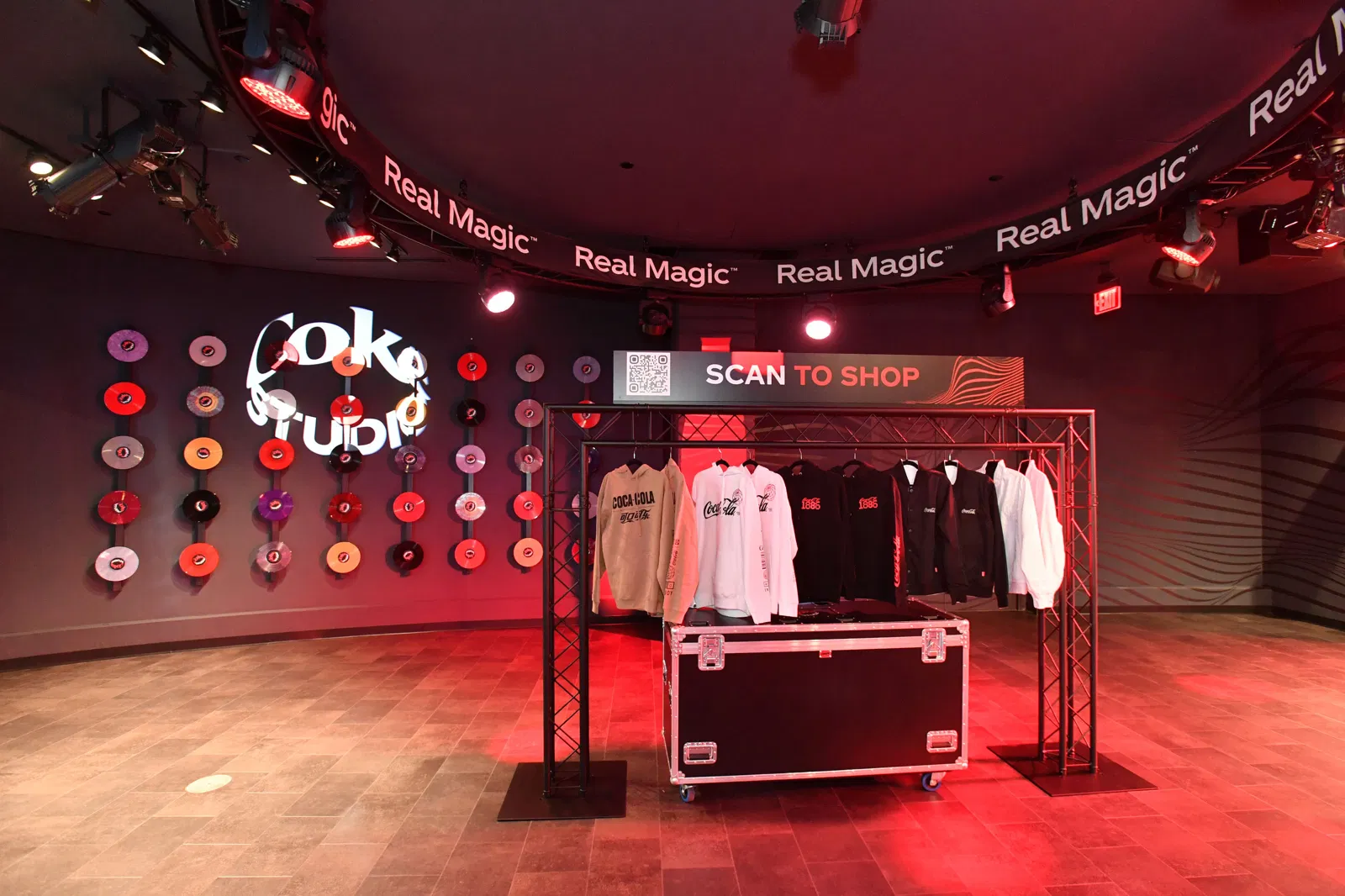 The red showroom with a musical theme. Vinyl records hang on the walls, the Coke Studio sign is illuminated, and in the center, under the spotlights, there is a display rack with branded clothing