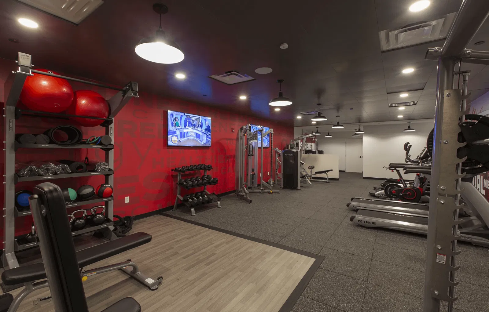 Compact gym room: a lot of balls and sports equipment. Red and white walls, with two TV on them.